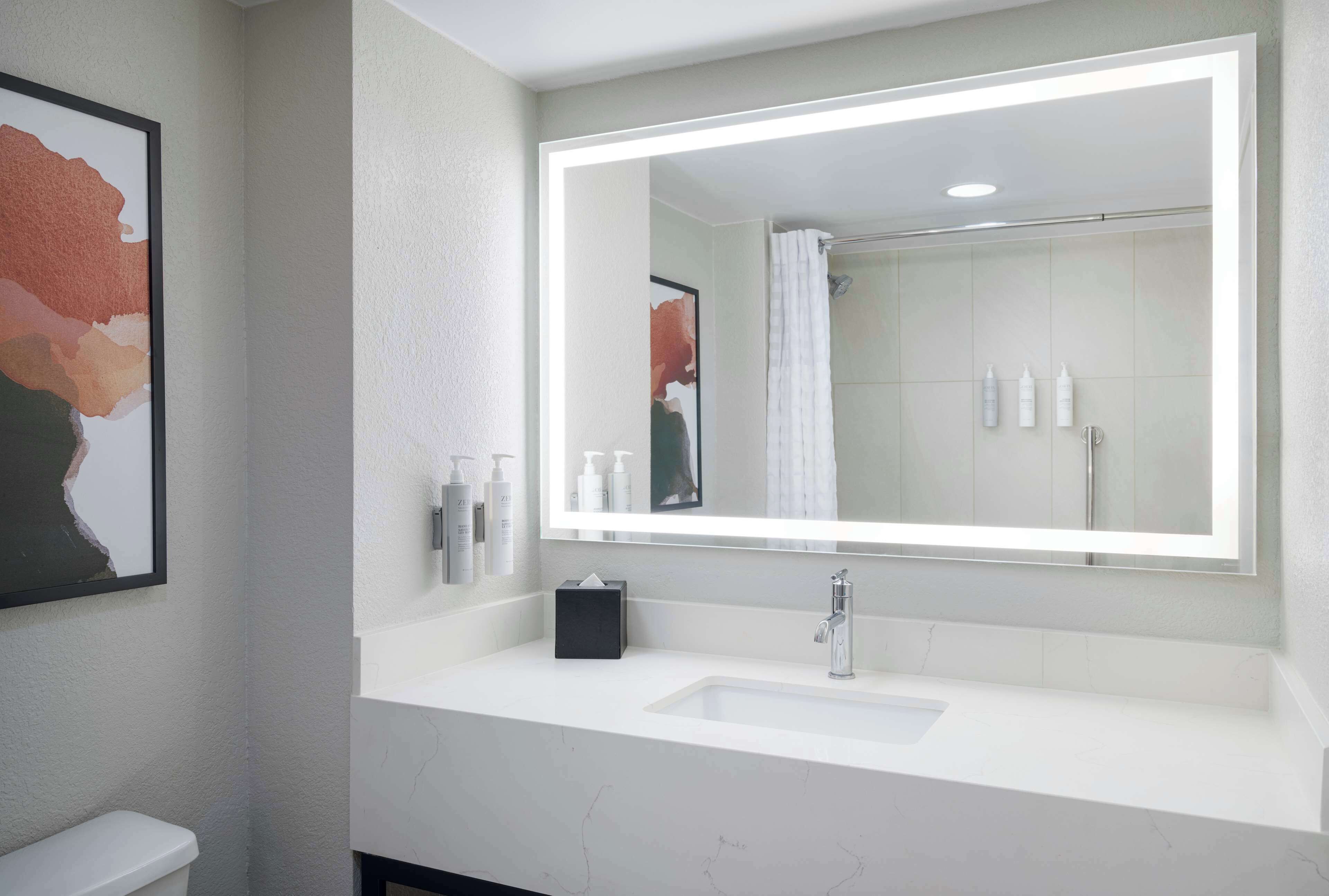 A modern bathroom with a large mirror, white sink, and a framed abstract painting on the wall.