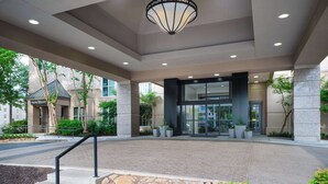 L’entrée moderne d’un hôtel, avec une grande porte en verre, un luminaire en métal noir et une zone pavée de briques agrémentée de plantes en pot.