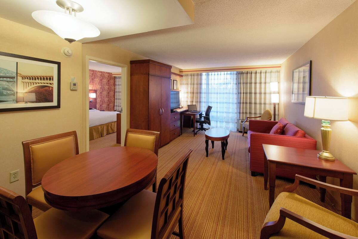 A hotel room with a dining table, chairs, a sofa, a desk with a chair, a television, and a bed.