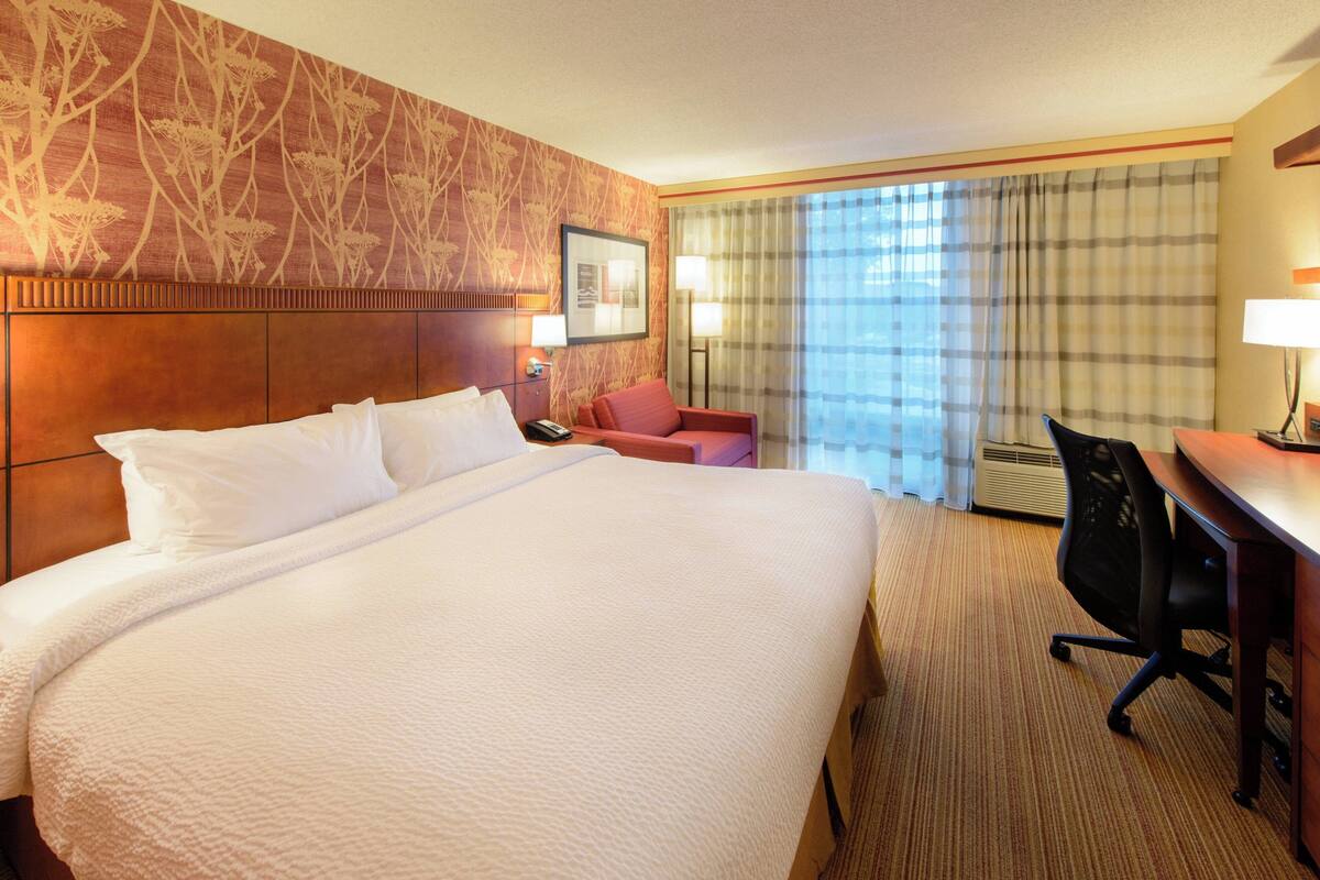 A hotel room with a large bed, a desk, a chair, a nightstand, a lamp, and a window with curtains.