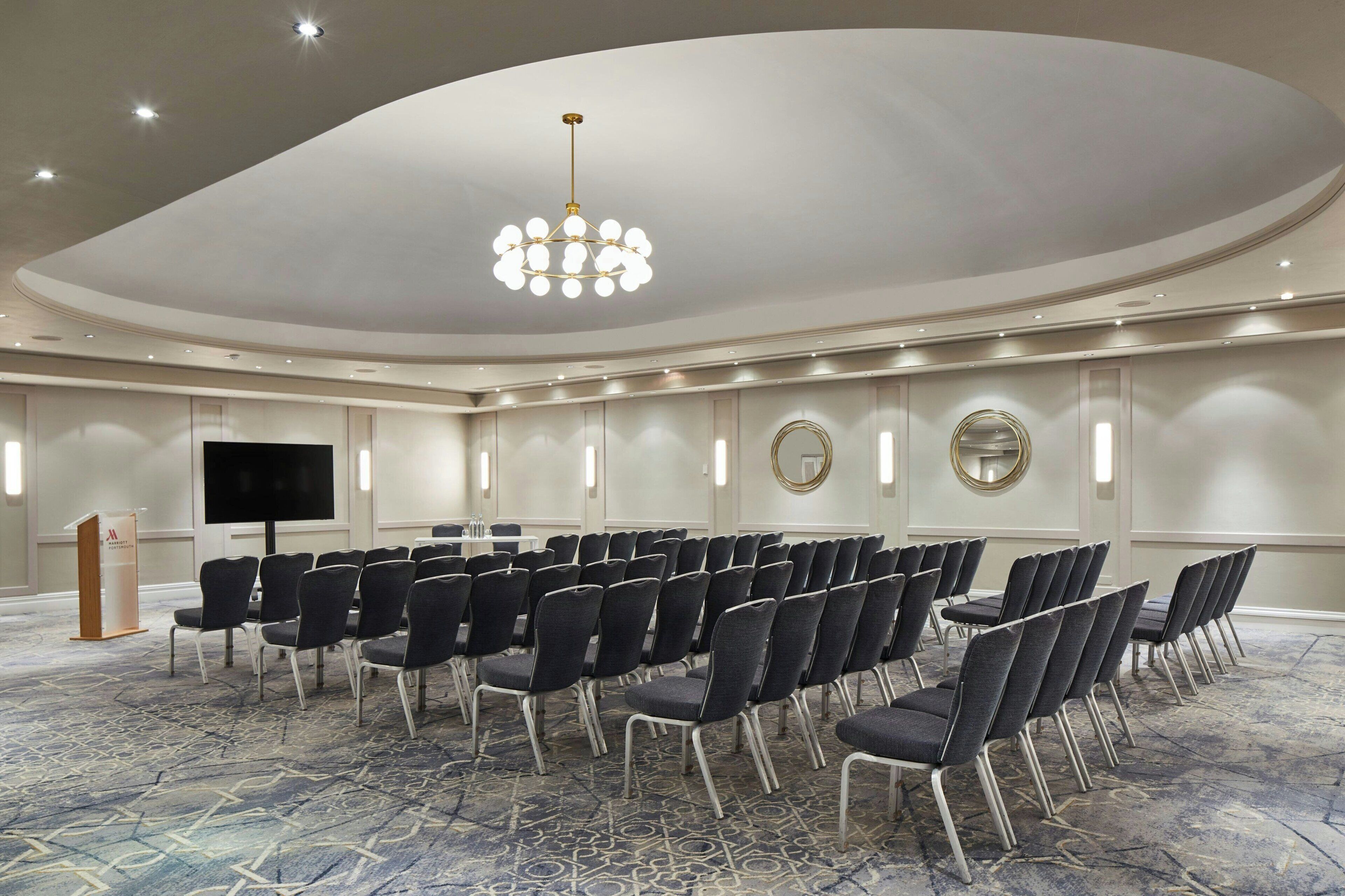 Een congreszaal met rijen stoelen, een spreekgestoelte, een groot scherm en een kroonluchter.