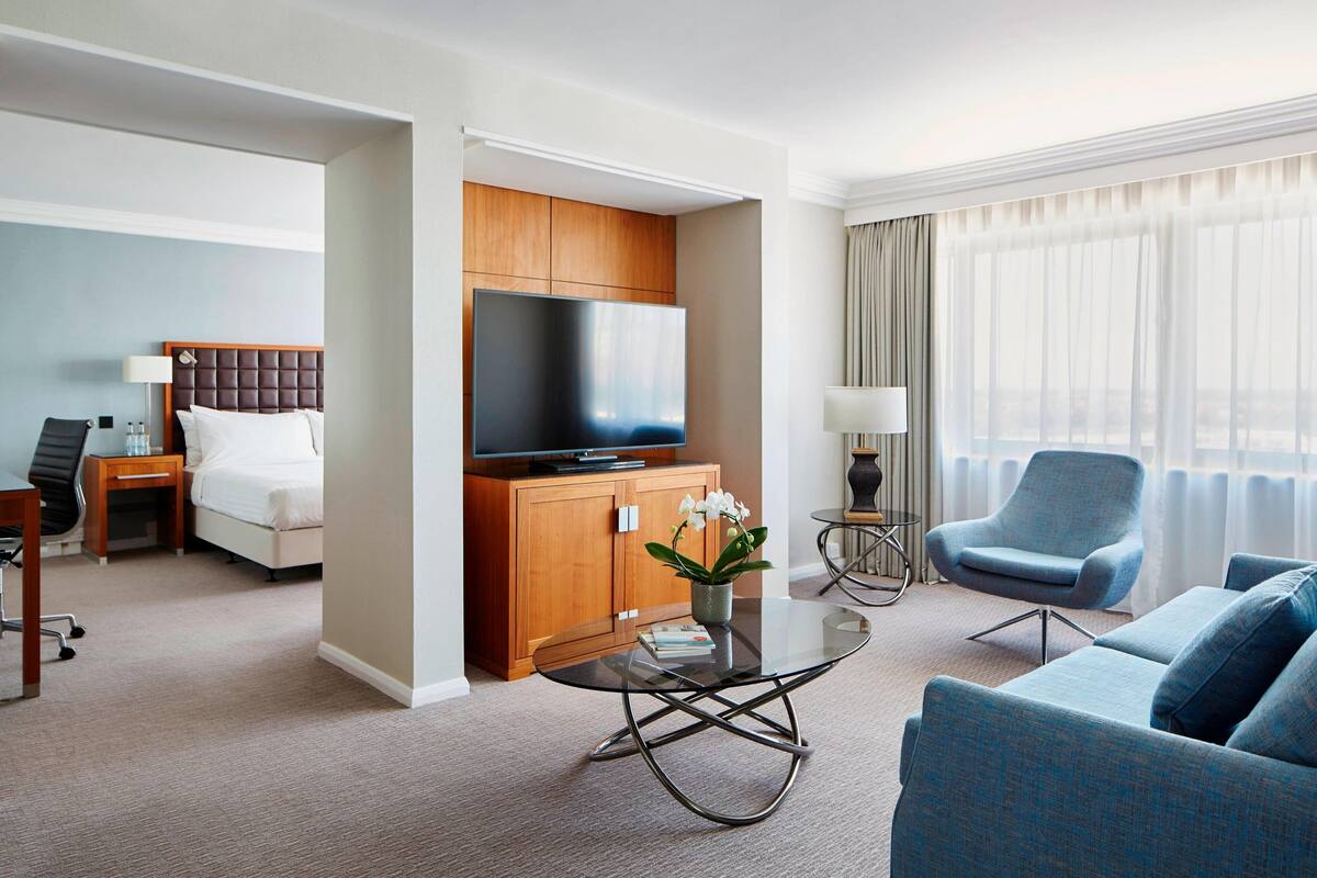 A modern hotel room with a bed, a TV, a sofa, a coffee table, and a view of the city through large windows.