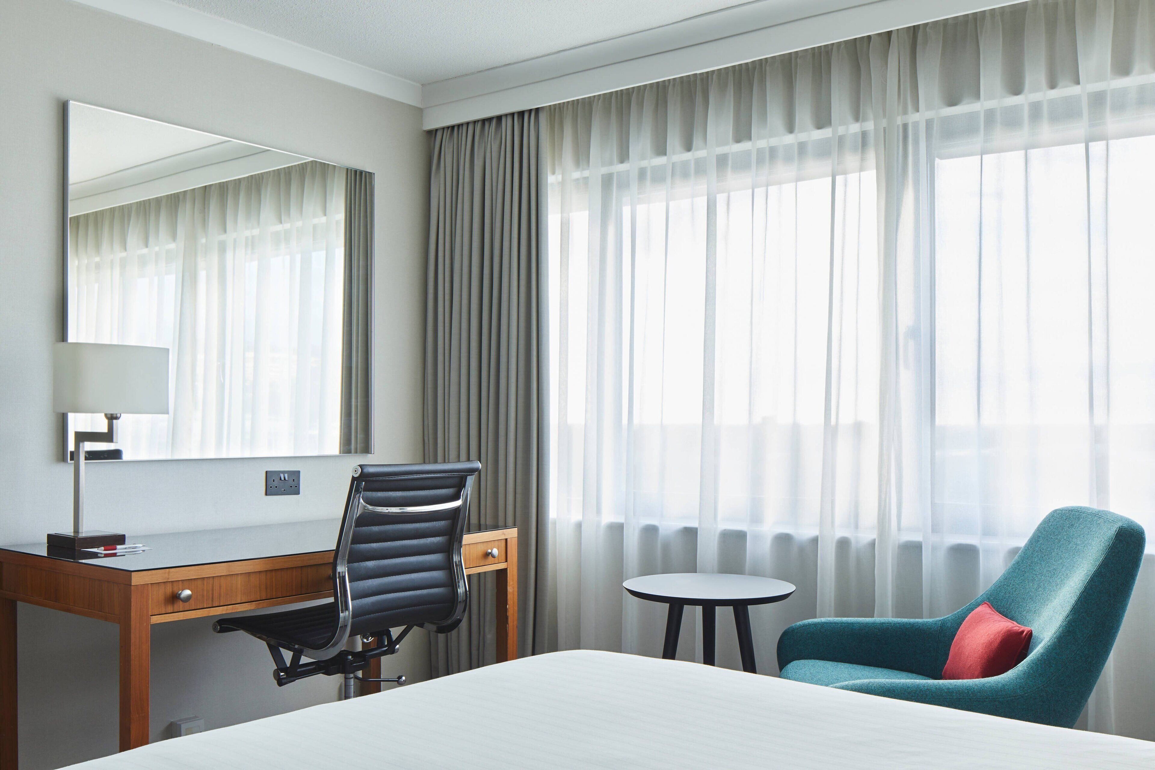 A hotel room with a bed, a desk with a chair, a small table, and a large window with curtains.