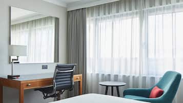 A hotel room with a bed, a desk with a chair, a small table, and a large window with curtains.