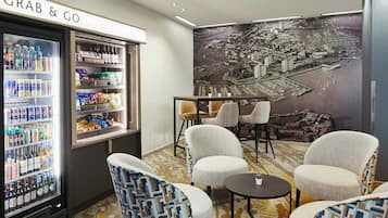 Habitación de hotel con refrigerador 'Grab & Go' provisto de bebidas, un gran mural con vista a la ciudad y una zona de estar con cuatro sillas y una mesita.