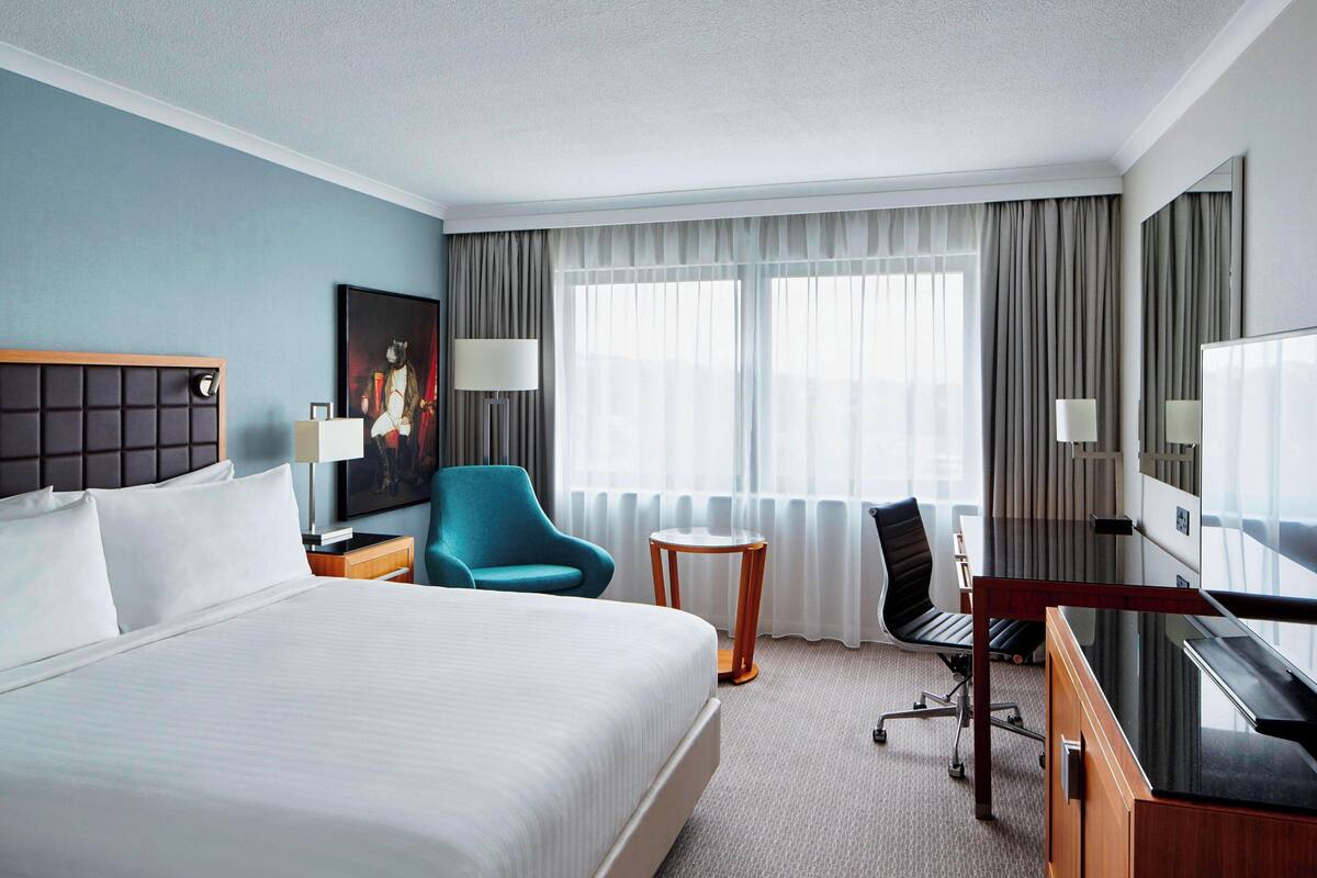 A hotel room with a large bed, a desk, a chair, a lamp, a TV, and a painting on the wall.
