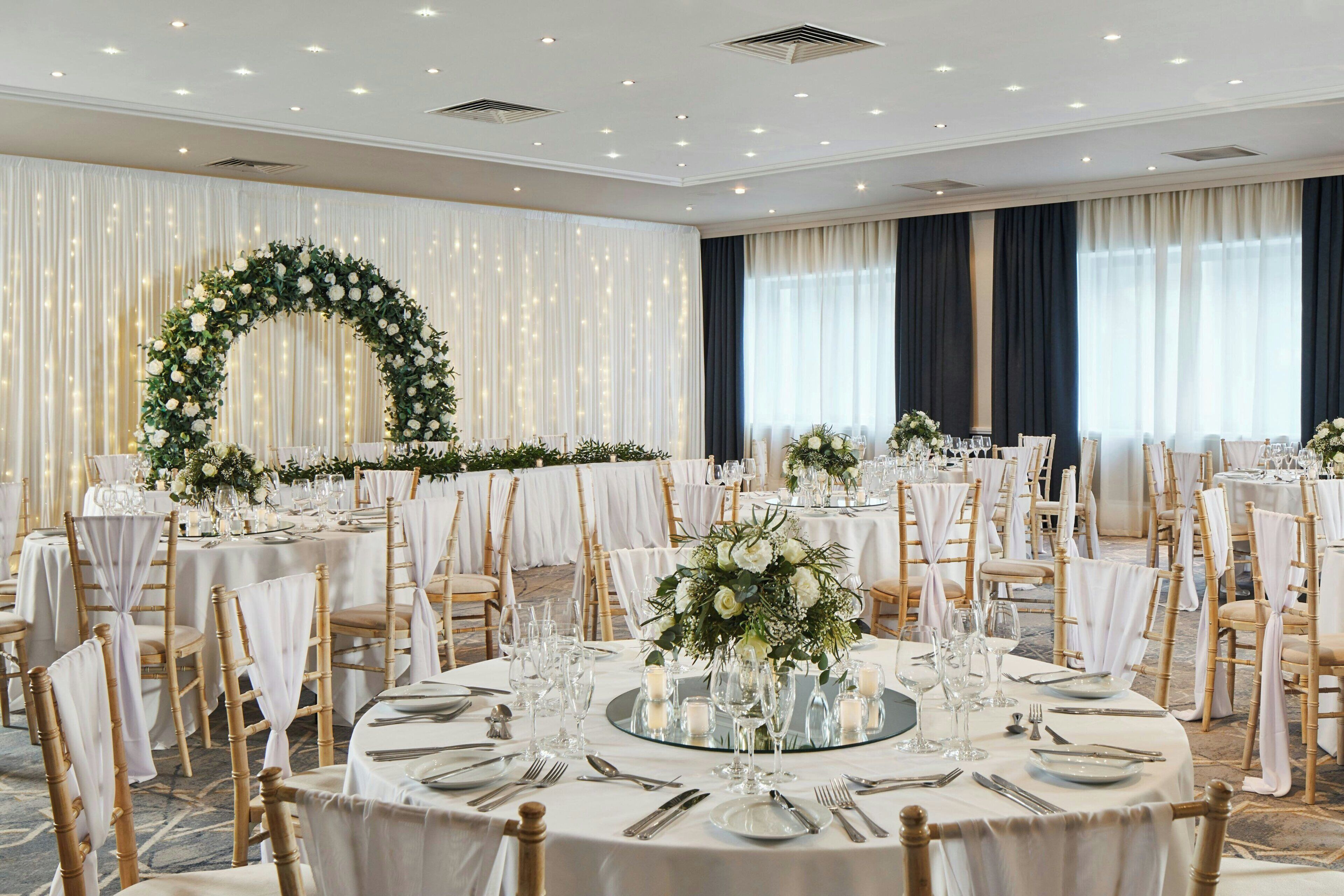 Un salón de banquetes con mesas redondas, manteles blancos y centros de mesa florales.