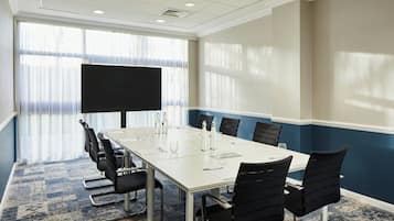 Un salón de conferencias con una larga mesa, sillas negras, una pantalla de proyector y un amplio ventanal con cortinas translúcidas.