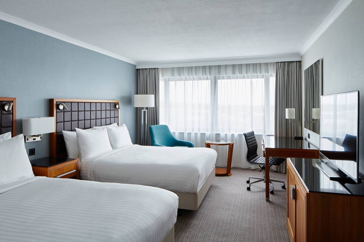 A hotel room with two beds, a desk, a chair, a TV, and a window with curtains.