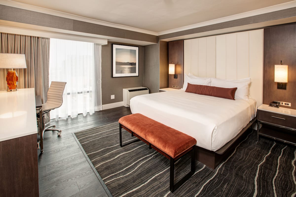A hotel room with a large bed, a bench, a desk with a chair, a desk lamp, a wall-mounted phone, and a window with curtains.