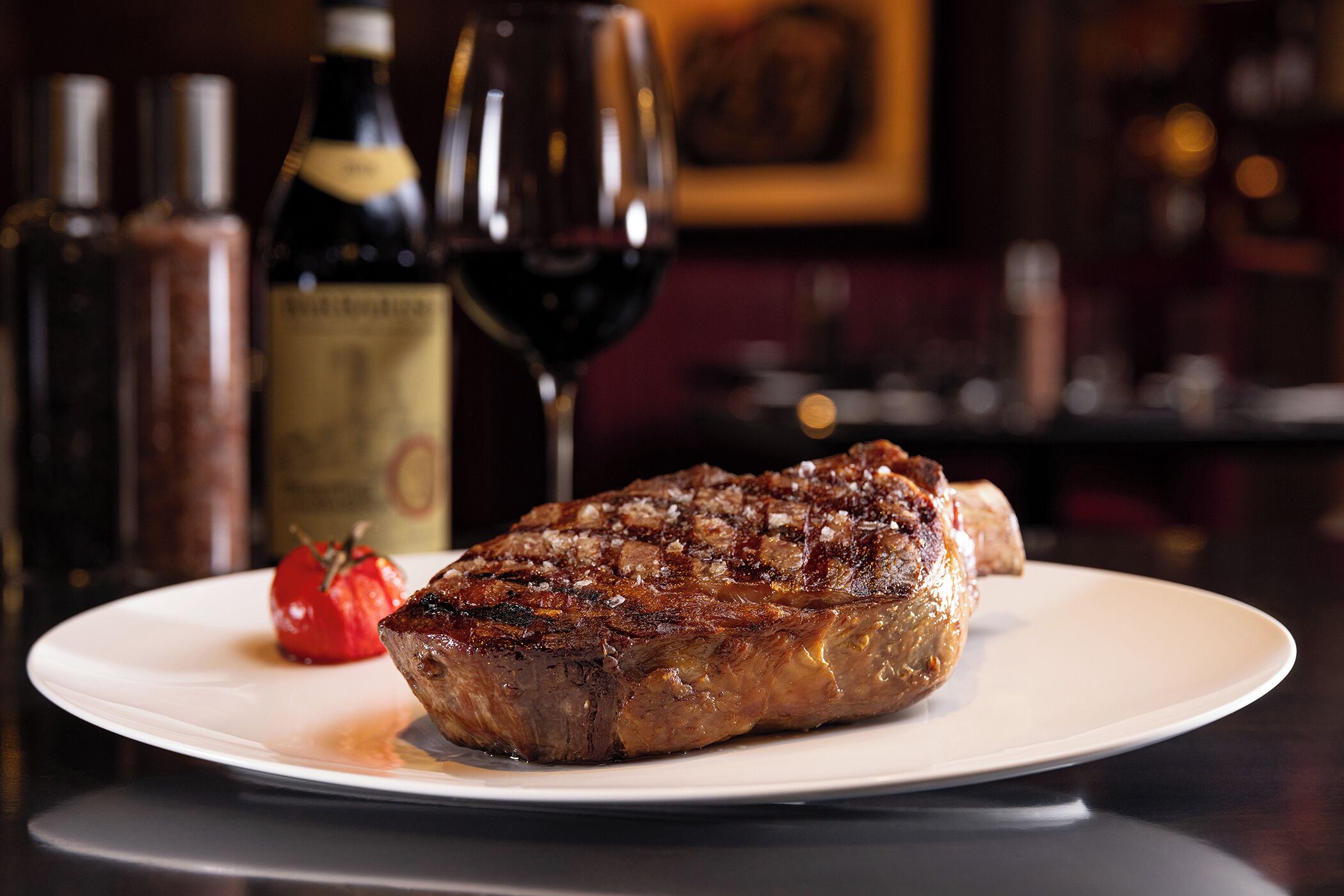 A steak on a plate with a glass of red wine and a bottle of wine in the background.