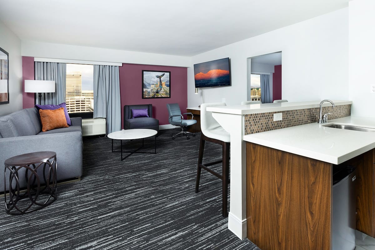 A modern hotel room with a grey sofa, a small round table, a desk with a sink, and a cityscape painting on the wall.