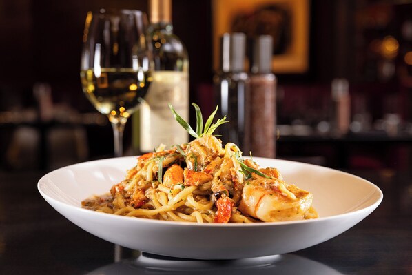 A plate of pasta with shrimp and vegetables, accompanied by a glass of white wine and a bottle of olive oil.
