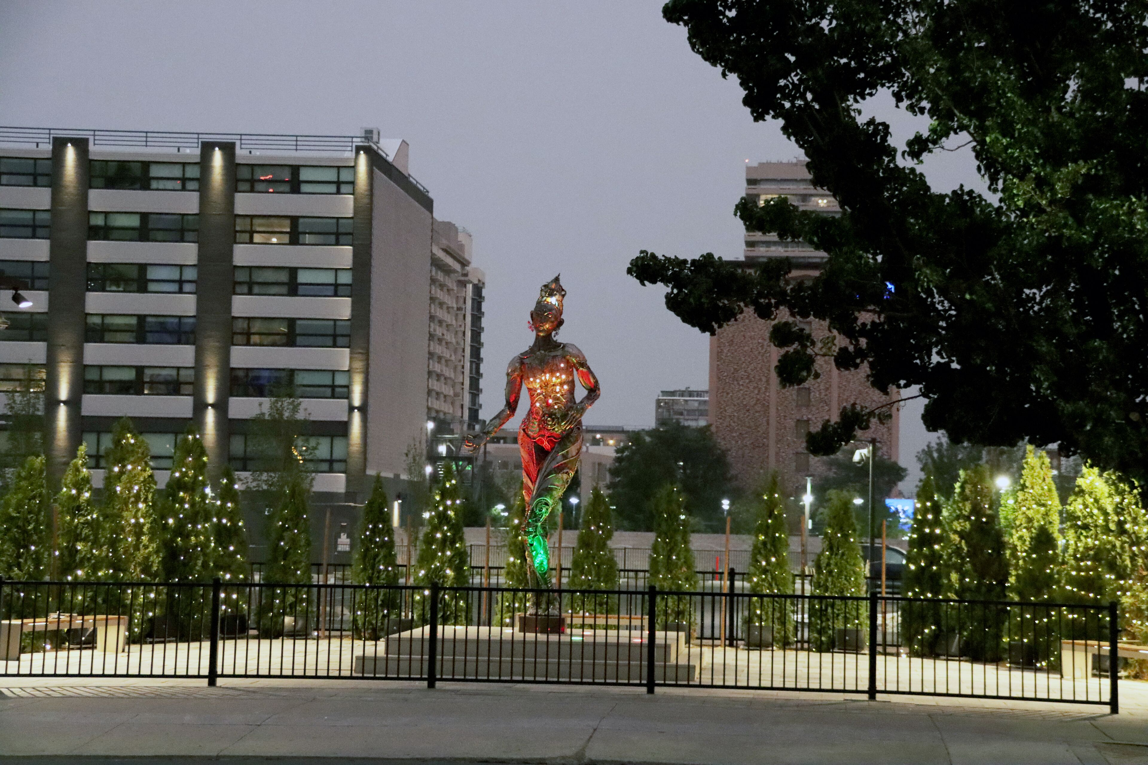 Una estatua en una plaza, rodeada de árboles y edificios, con una cerca alrededor.