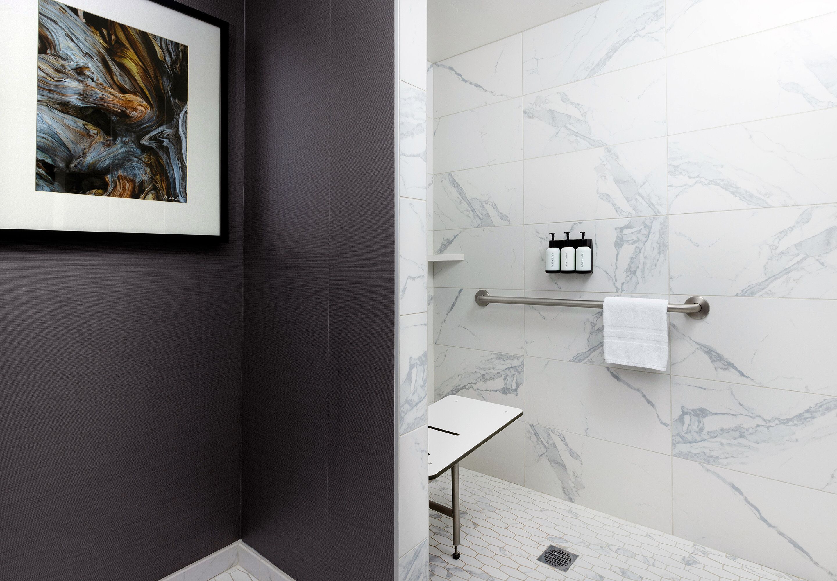 A modern bathroom with marble tiles, a framed artwork, a towel rack, and a small table.