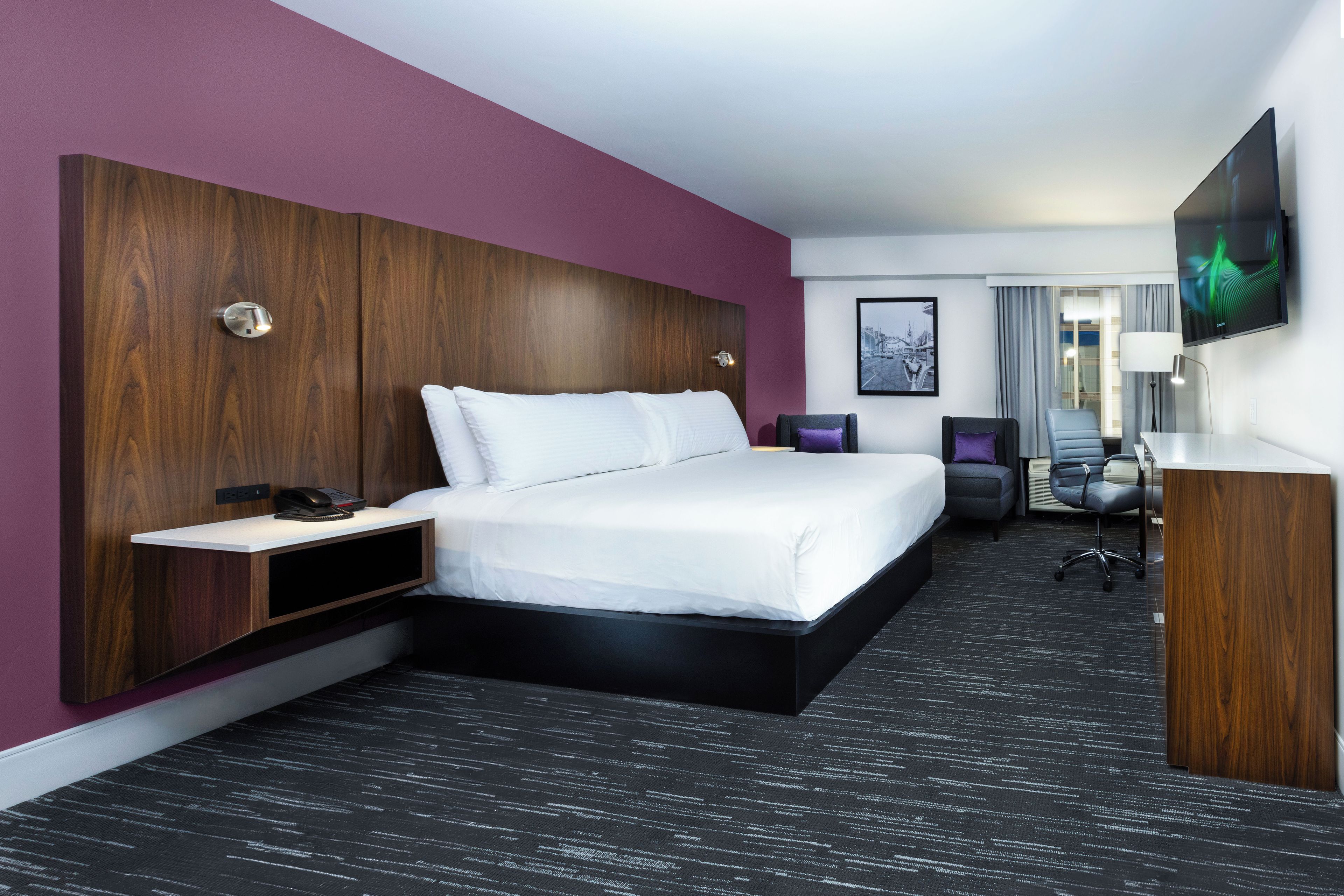 Ein Hotelzimmer mit einem großen Bett, einem Schreibtisch, einem Stuhl, einem Fernseher und einem Fenster mit Vorhängen.