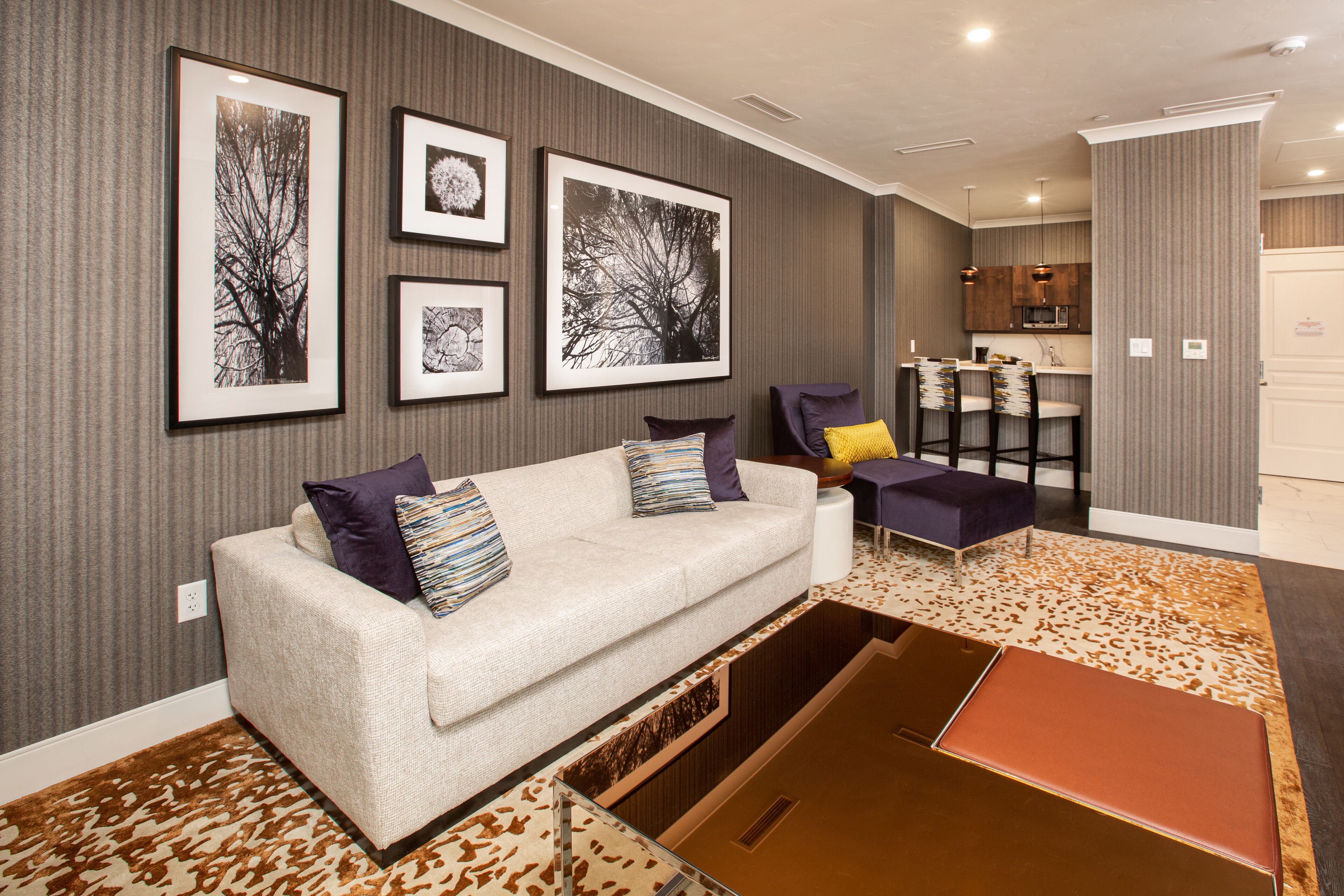 A modern living room with a sofa, chairs, and a coffee table. There are framed pictures on the wall and a kitchen area in the background.