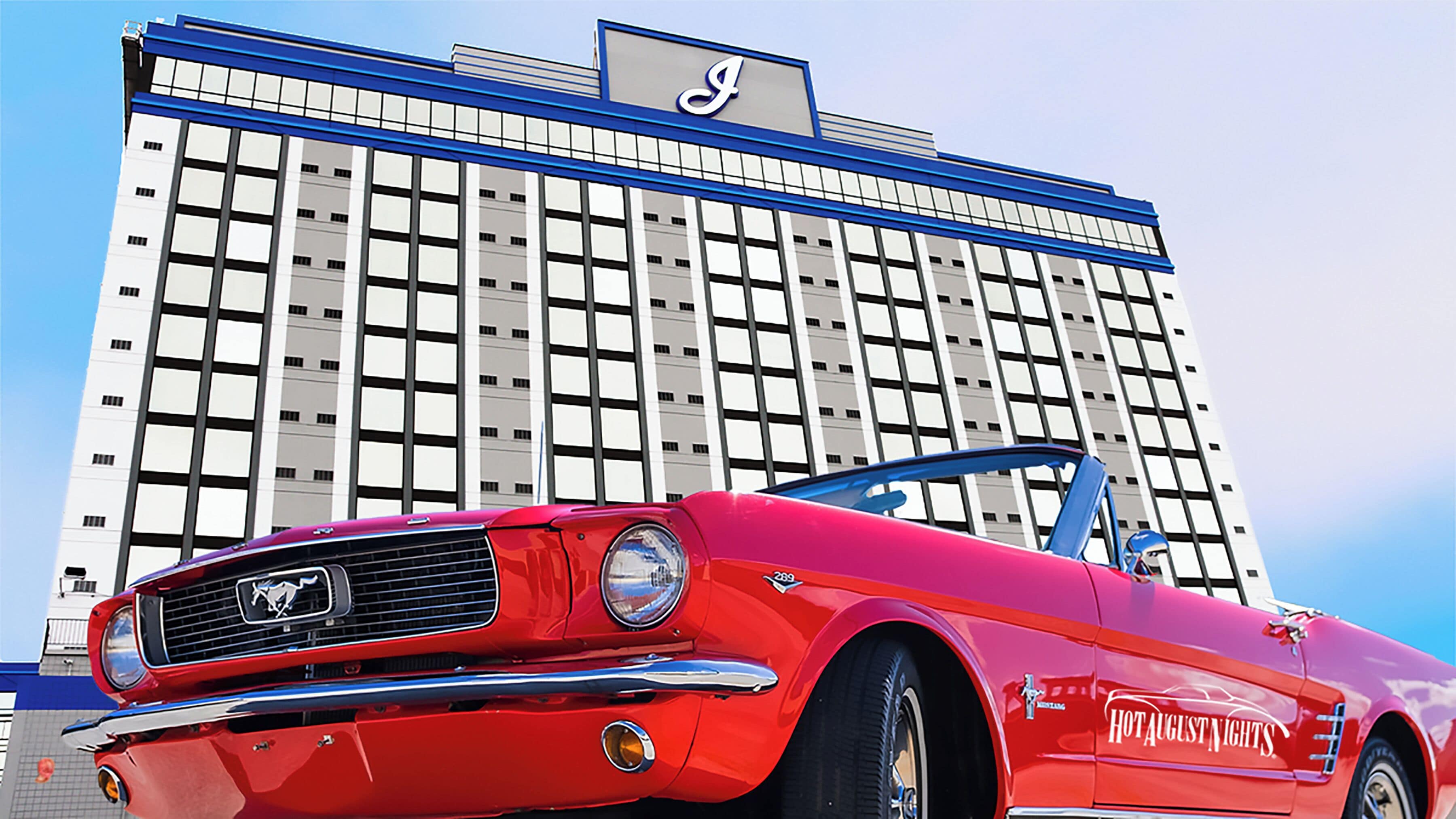 Un coche clásico rojo estacionado frente a un hotel con un llamativo letrero con la letra 'J'.