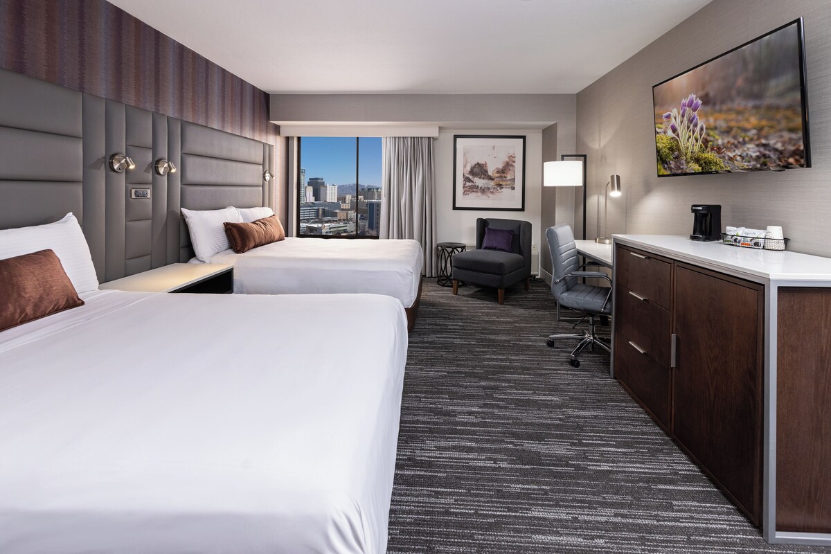 A hotel room with two beds, a desk, a chair, a coffee maker, and a city view.