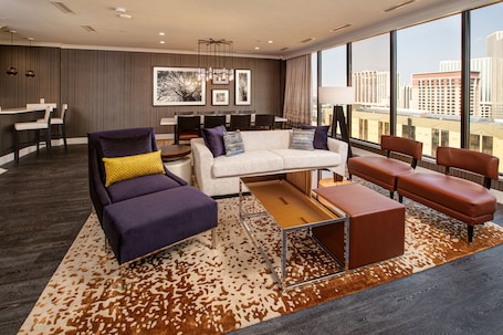 A modern hotel room with a sofa, armchairs, a dining area, and large windows offering a city view.