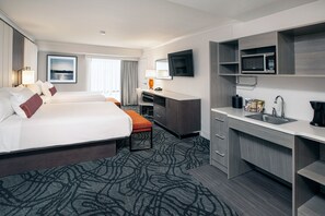 A hotel room with two beds, a desk, a microwave, a sink, and a large window with a view.