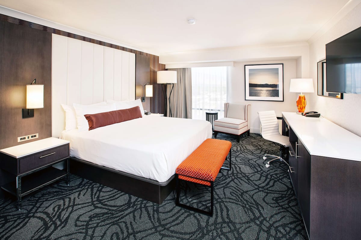 Une chambre d’hôtel avec un grand lit, des tables de chevet, un bureau, une chaise, un banc, une télévision et une fenêtre avec des rideaux.