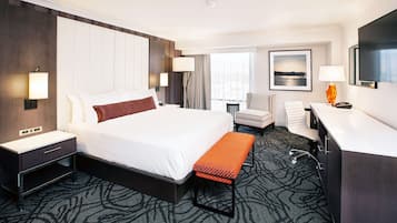 A hotel room with a large bed, bedside tables, a desk, a chair, a bench, a TV, and a window with curtains.
