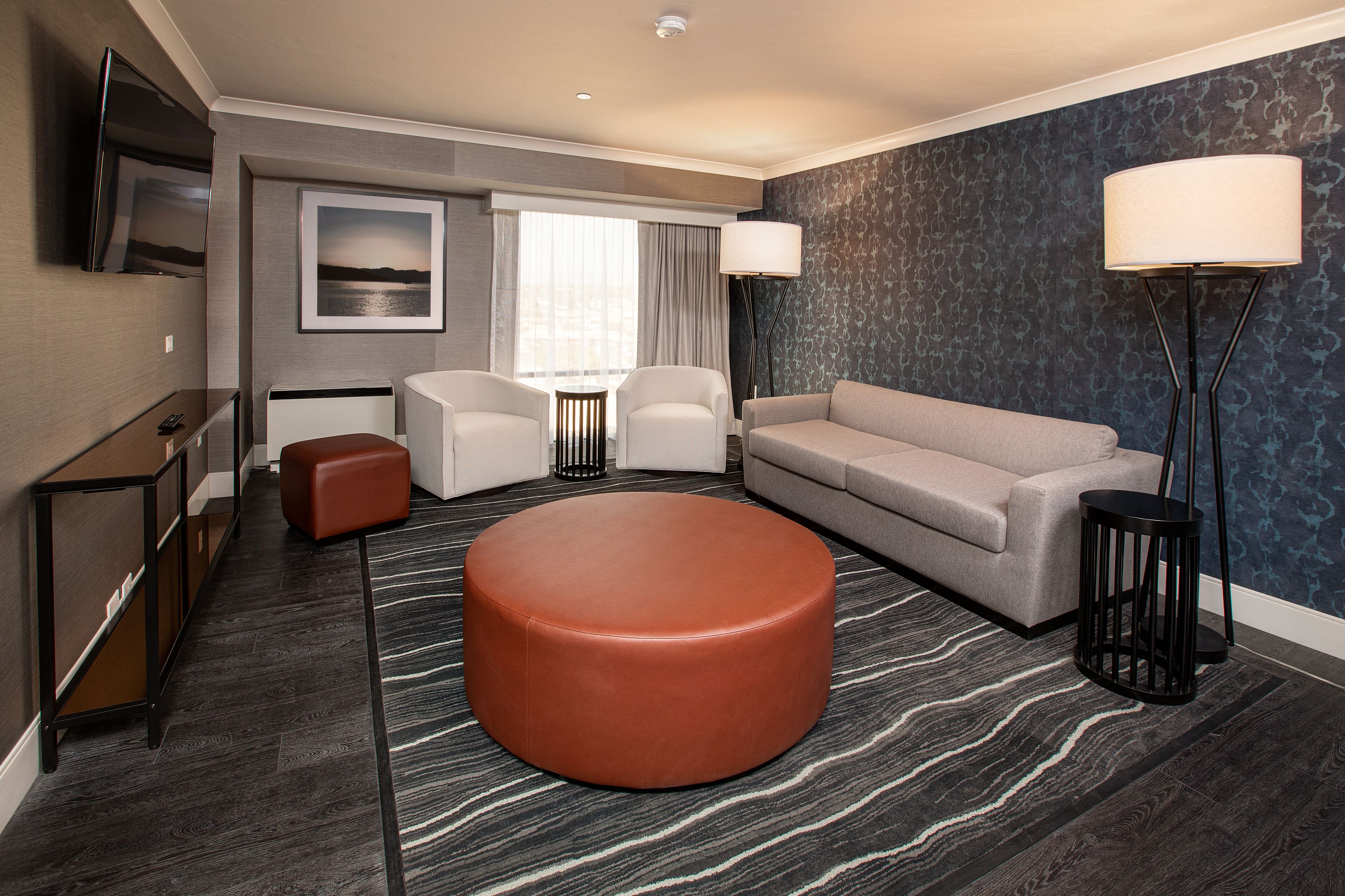 A modern hotel room with a sofa, ottoman, chairs, a TV, and a large window with curtains.