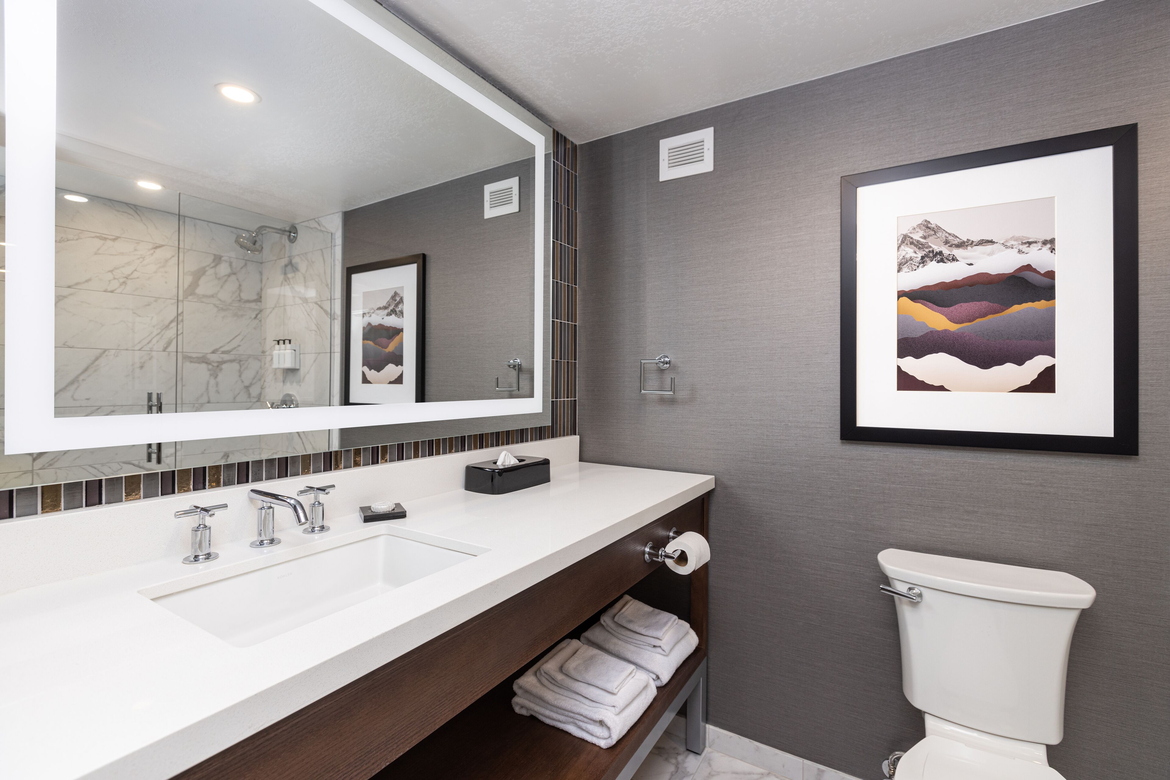 A modern bathroom with a white sink, a large mirror, a framed artwork on the wall, and a toilet.