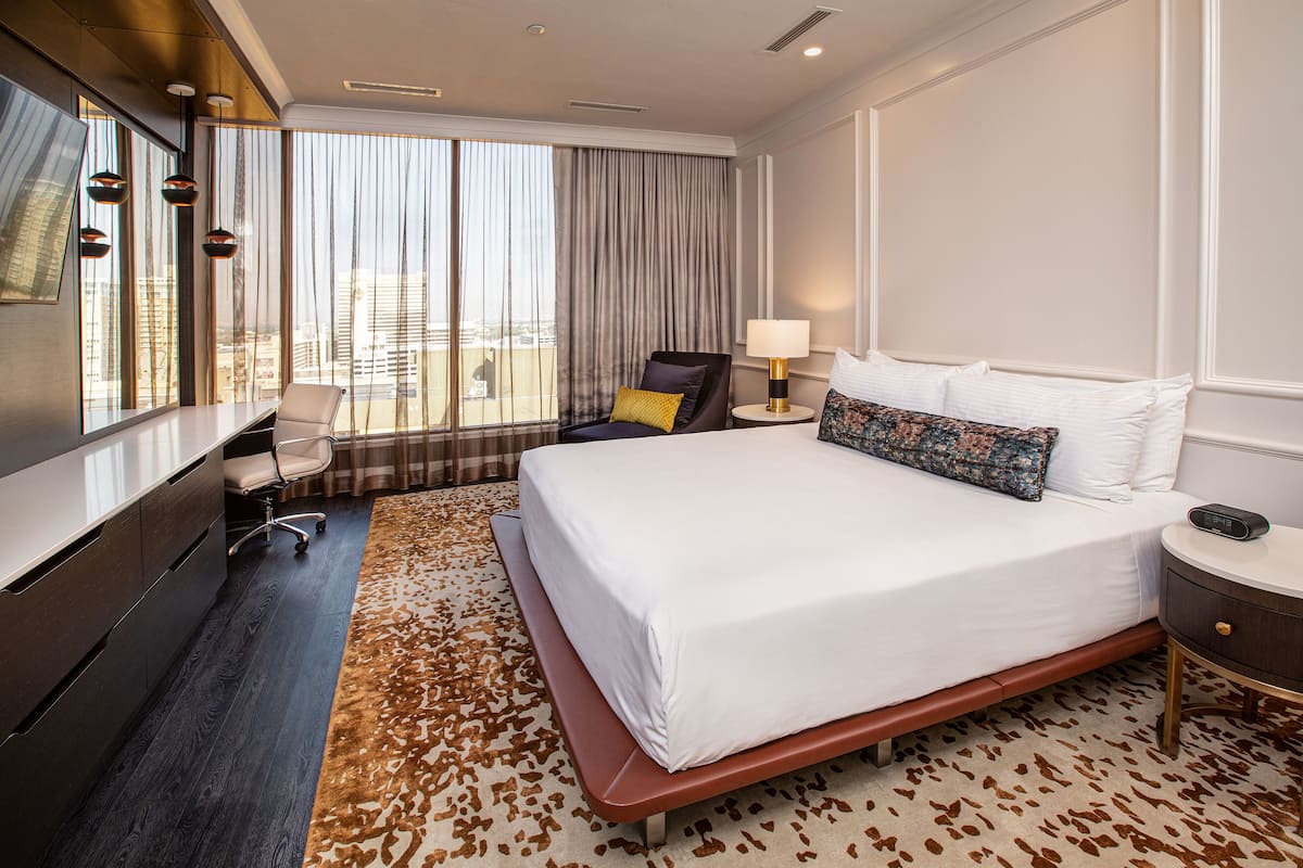 A hotel room with a large bed, a desk, a chair, a lamp, and a view of the city through large windows.
