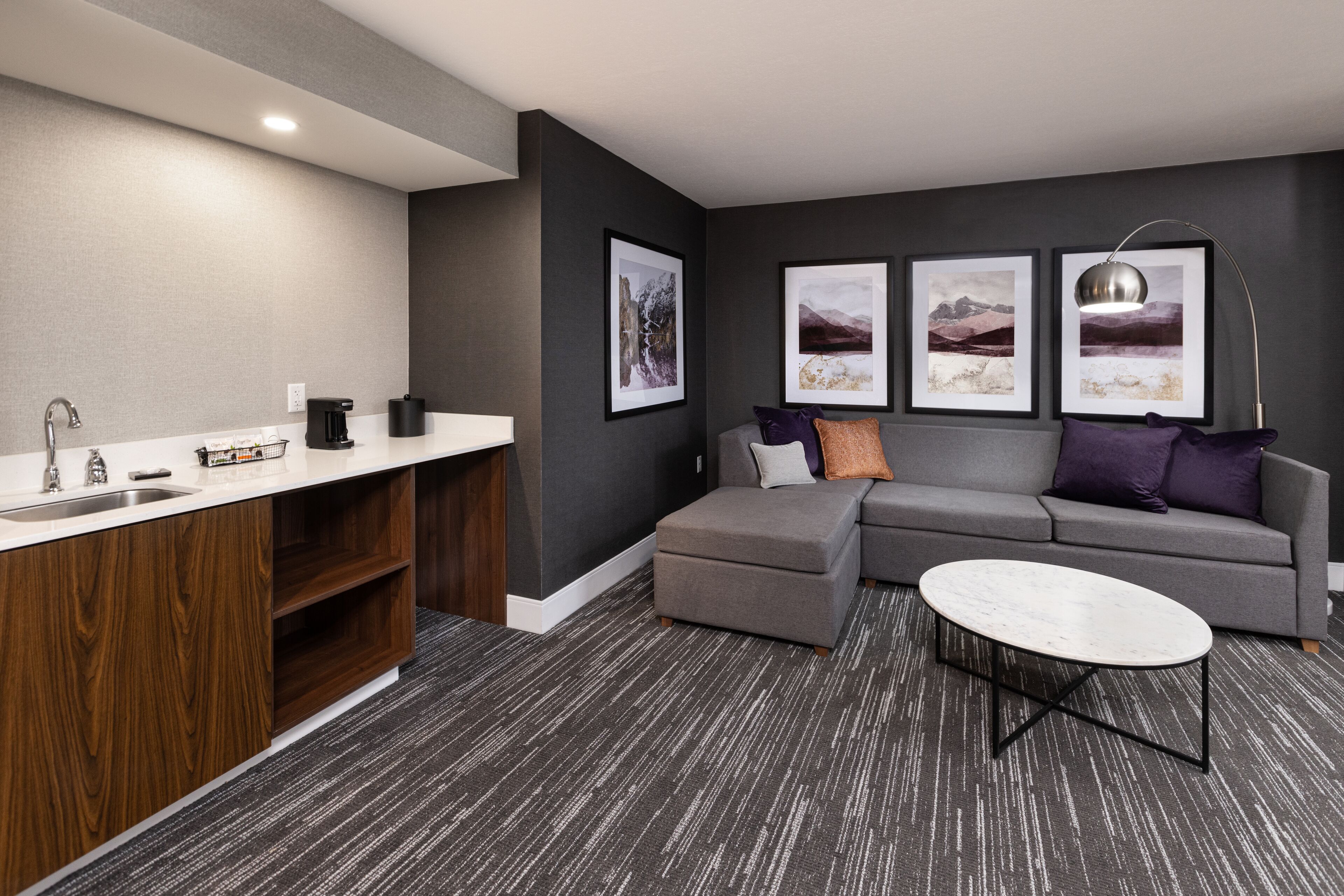 A modern living room with a grey sofa, a small coffee table, and a kitchen area with a sink and cabinets.