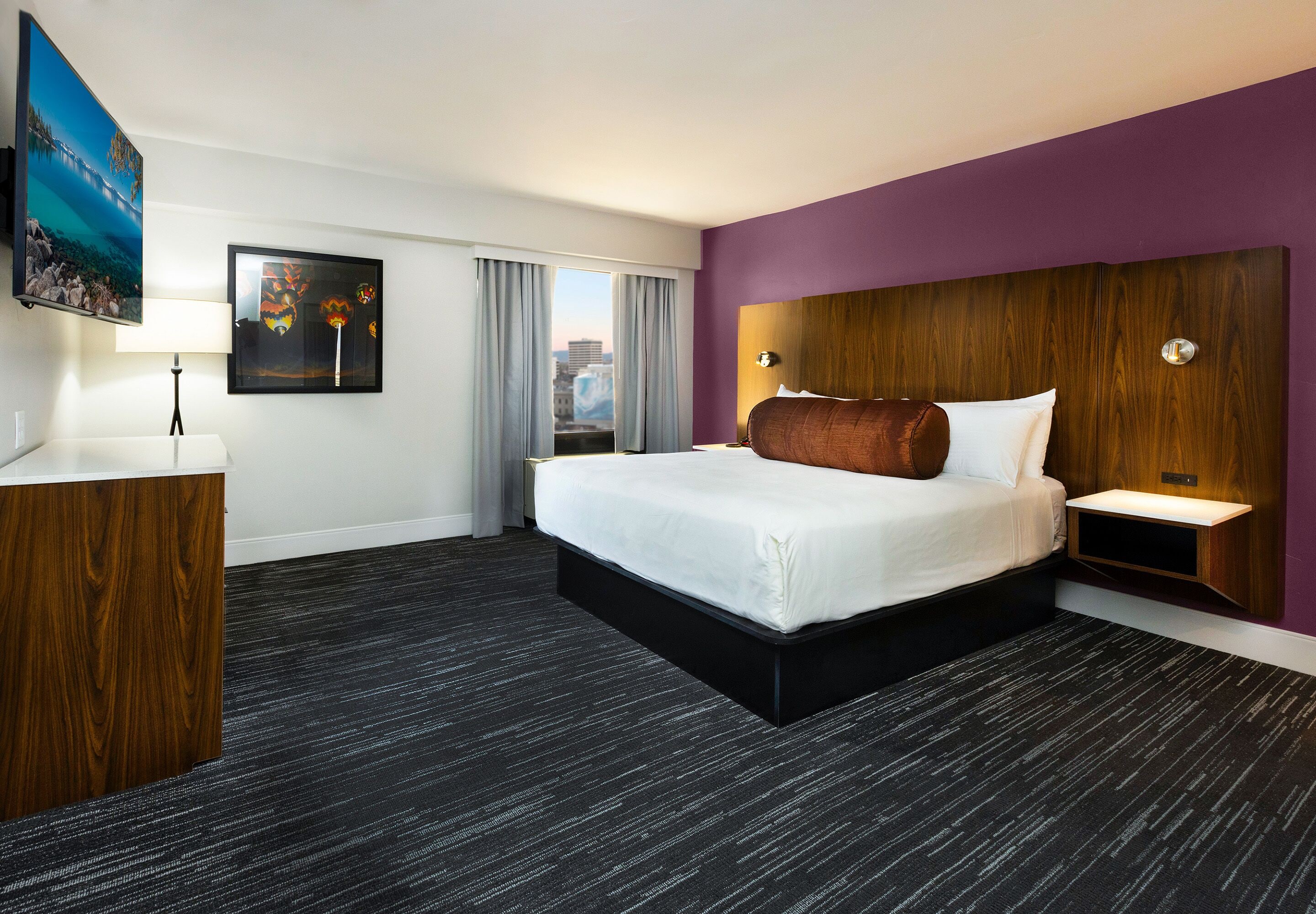 A hotel room with a large bed, a wooden headboard, a desk with a lamp, and a city view through the window.