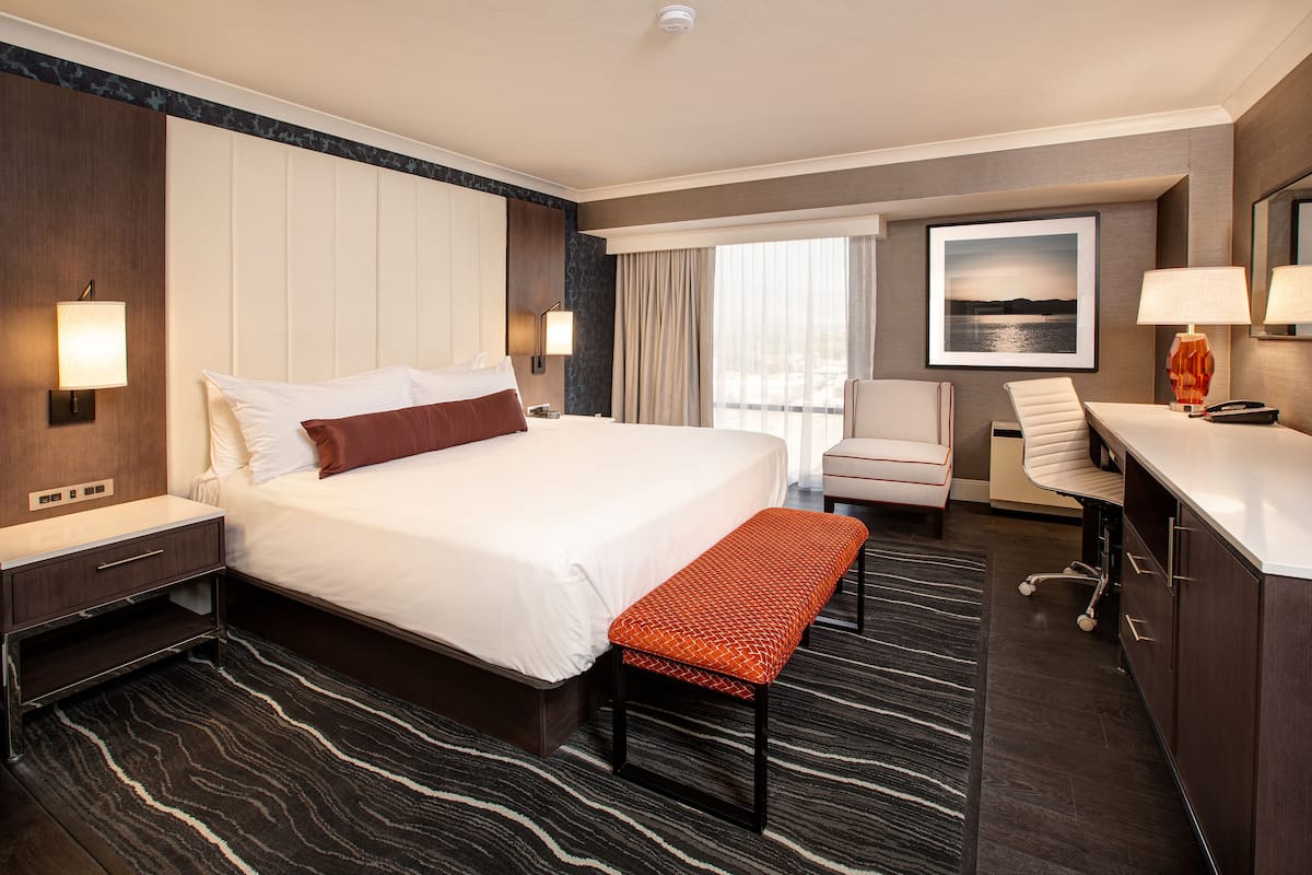 A hotel room with a large bed, a desk with a lamp, a chair, and a view of the outdoors.