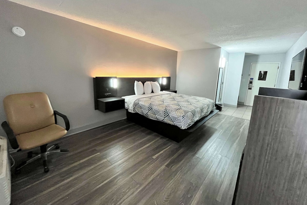 Una habitación de hotel moderna con cama, una silla, una mesita de noche y cabecero con iluminación integrada.