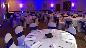 Un salón de banquetes con mesas redondas dispuestas para comer, sillas blancas con cintas azules e iluminación ambiental púrpura.