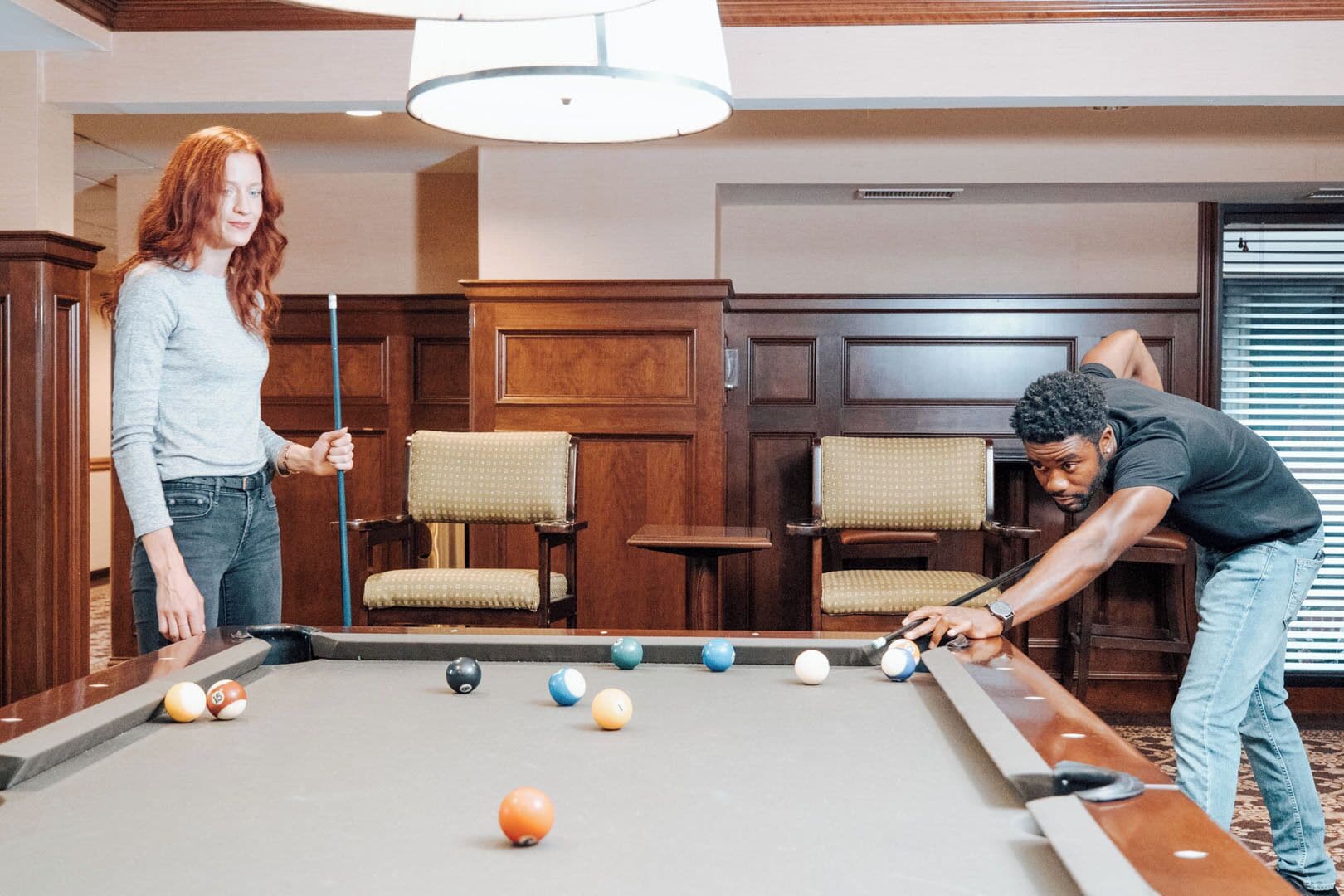 A pool table in a room with wooden paneled walls and two upholstered chairs.
