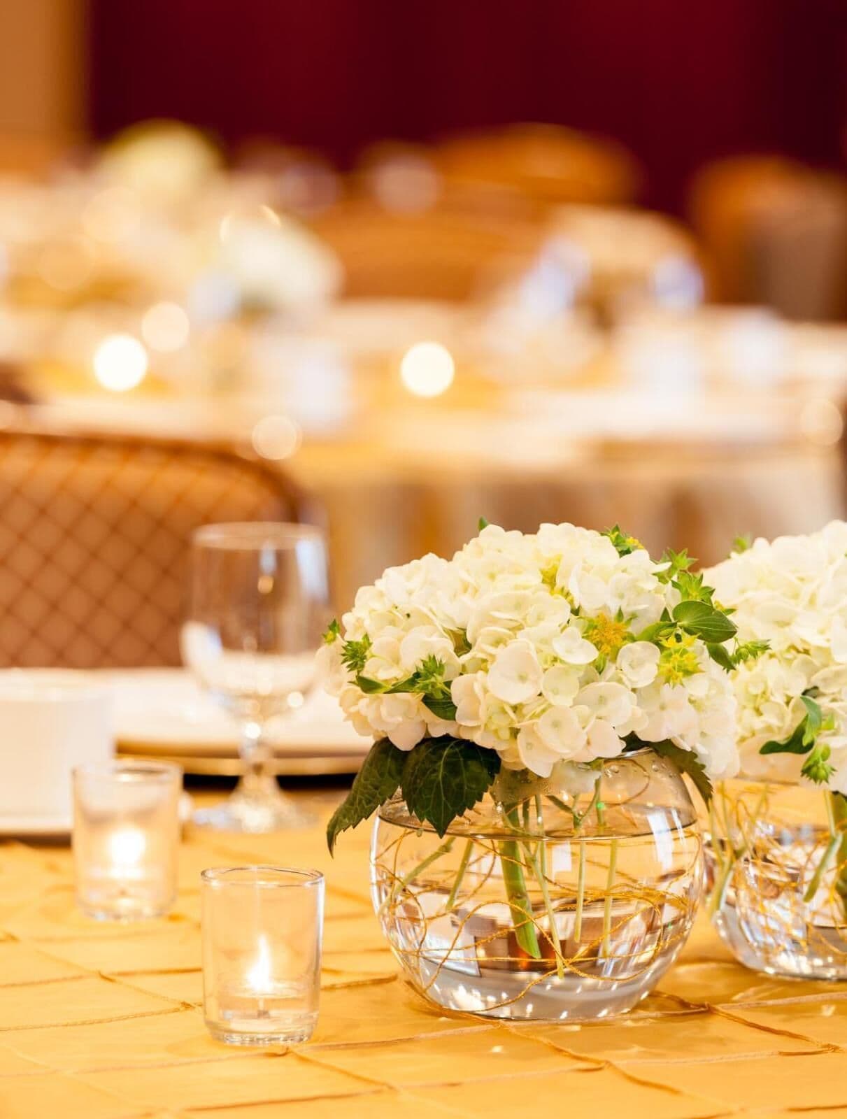 Een eettafel met een transparante glazen vaas gevuld met witte bloemen, een brandende kaars en een glas water.