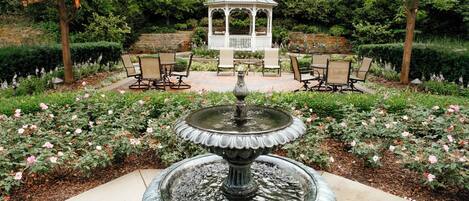 Un jardin avec une fontaine centrale, entouré de rosiers et agrémenté d’un belvédère.