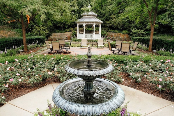 Un jardín con una fuente central, rodeado de rosales y un pabellón.