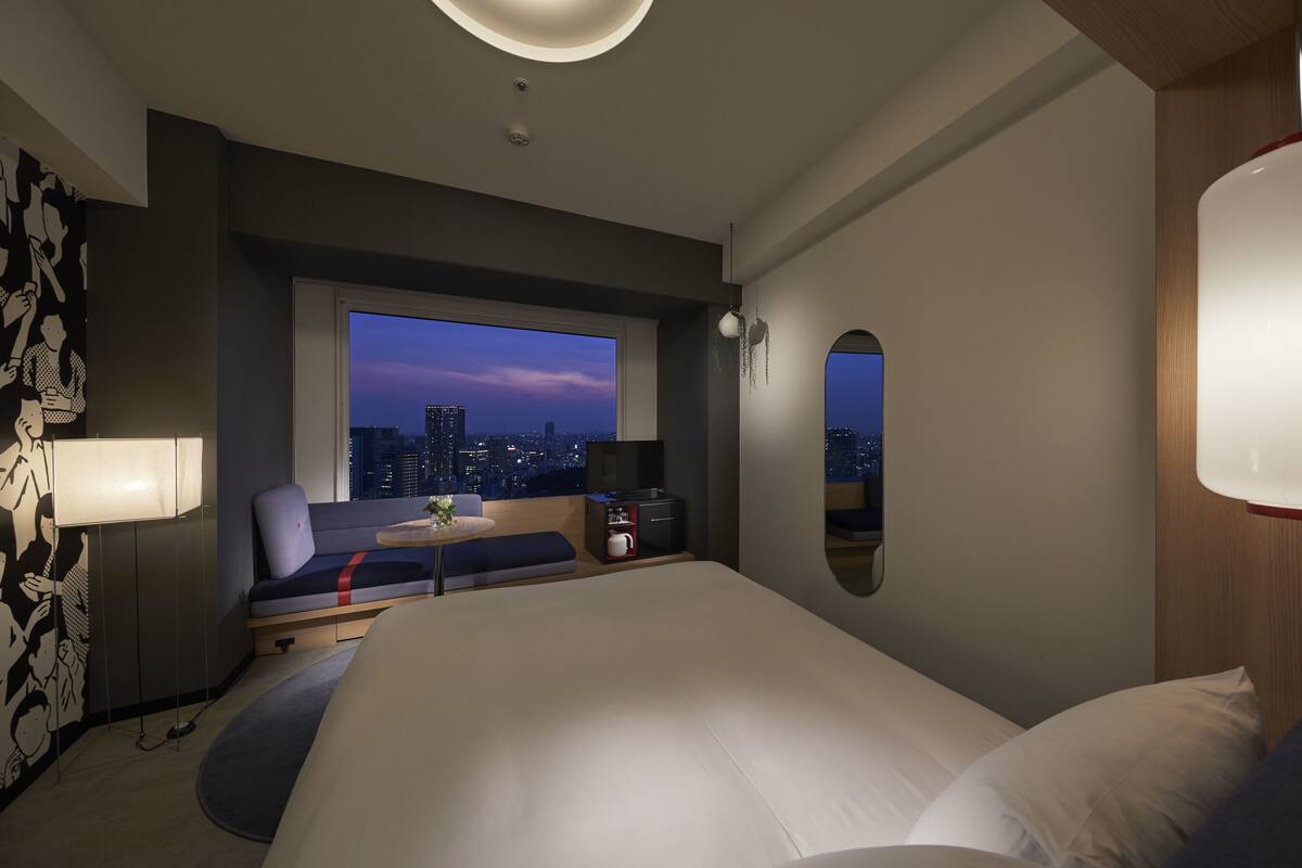 A modern hotel room with a large bed, a seating area with a view of the cityscape, a desk with a lamp, and a large mirror.