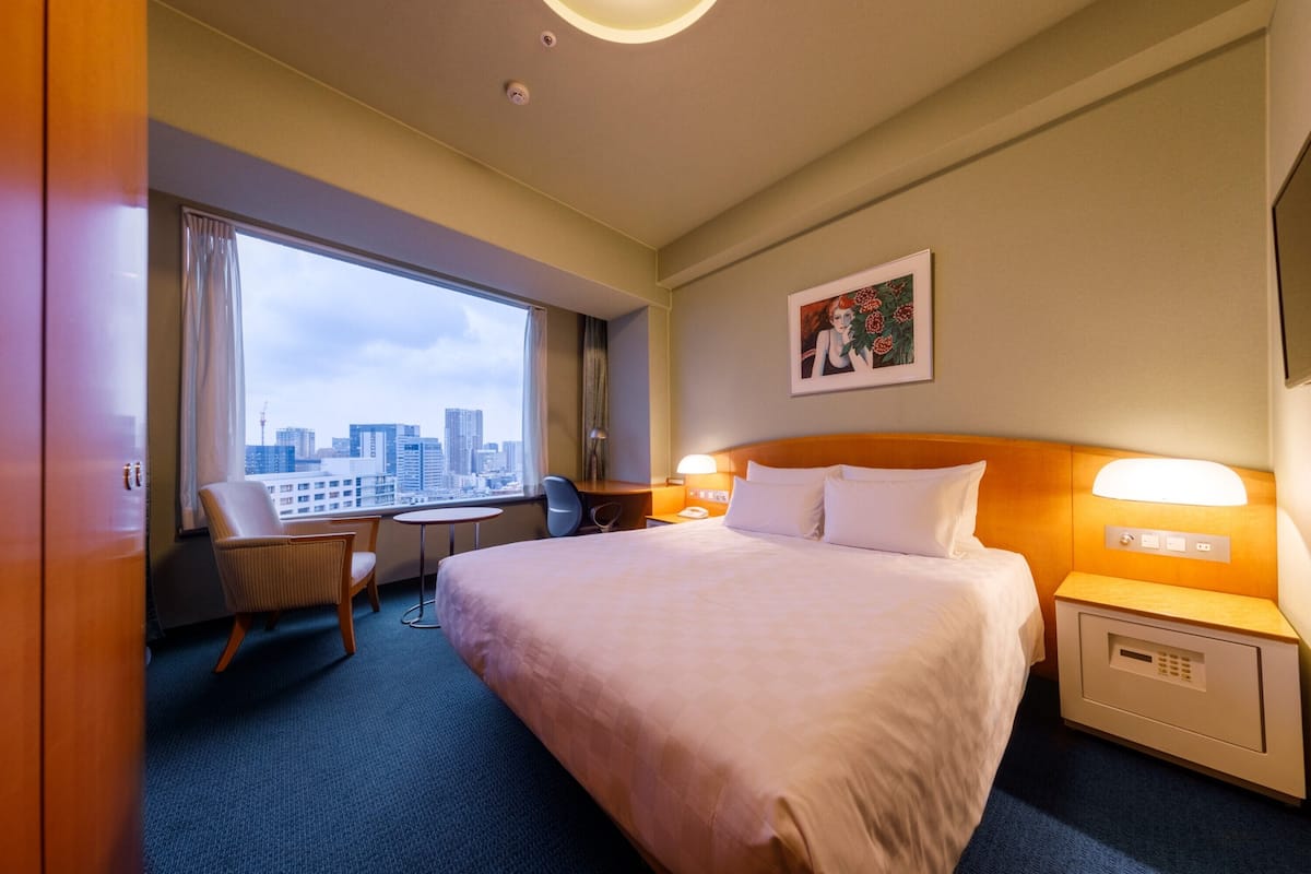 A hotel room with a large bed, a desk, a chair, and a window with a city view.