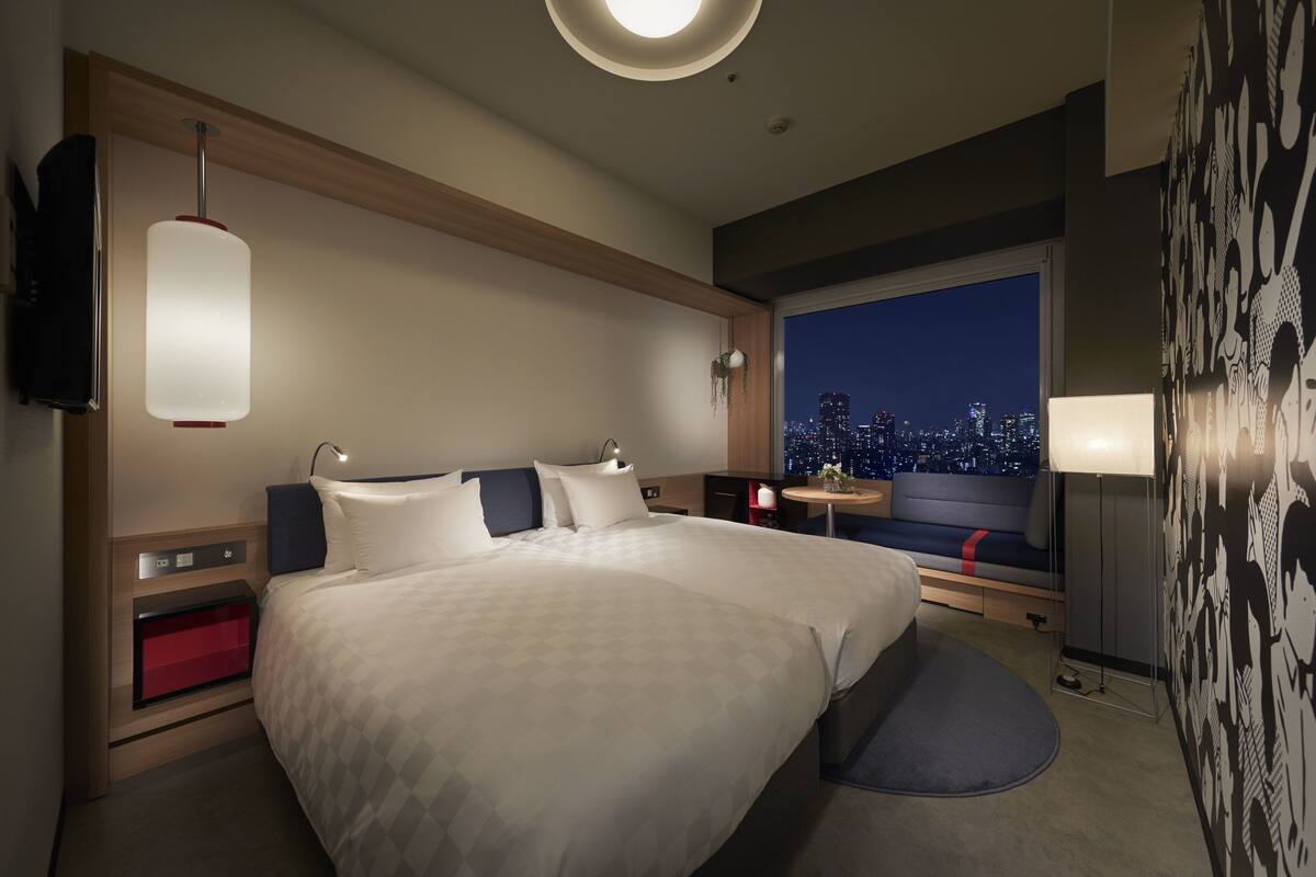 A modern hotel room with a large bed, bedside tables, a TV, a city view, and a small seating area by the window.