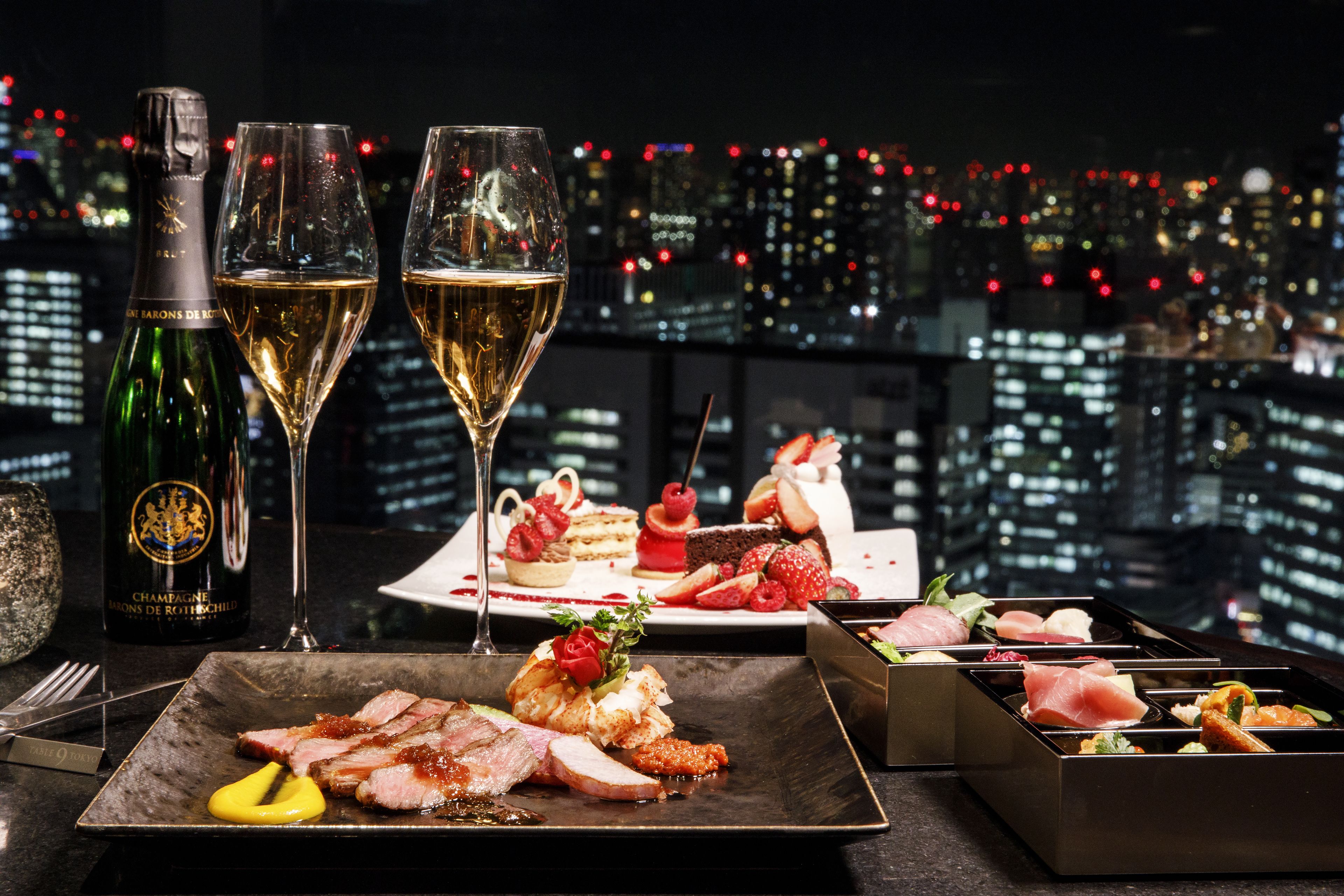A dining table with a bottle of champagne, two glasses, and a variety of desserts and appetizers.