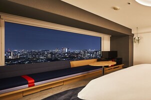 A hotel room with a large window offering a city view at night, a wooden bench with cushions, a small round table, and a bed.