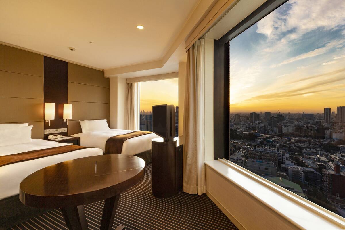 A hotel room with two beds, a TV, a desk, and a large window with a city view.