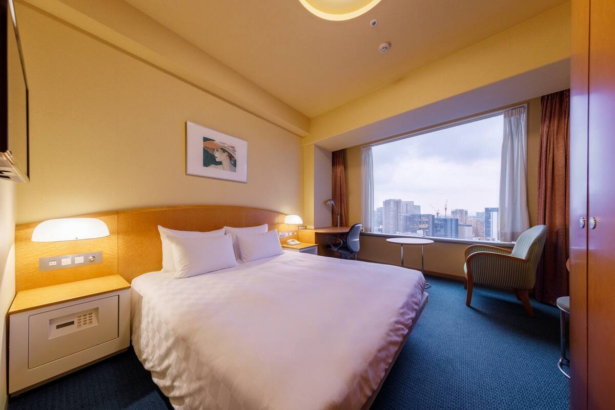 A hotel room with a large bed, a desk, a chair, a small table, and a window with a city view.