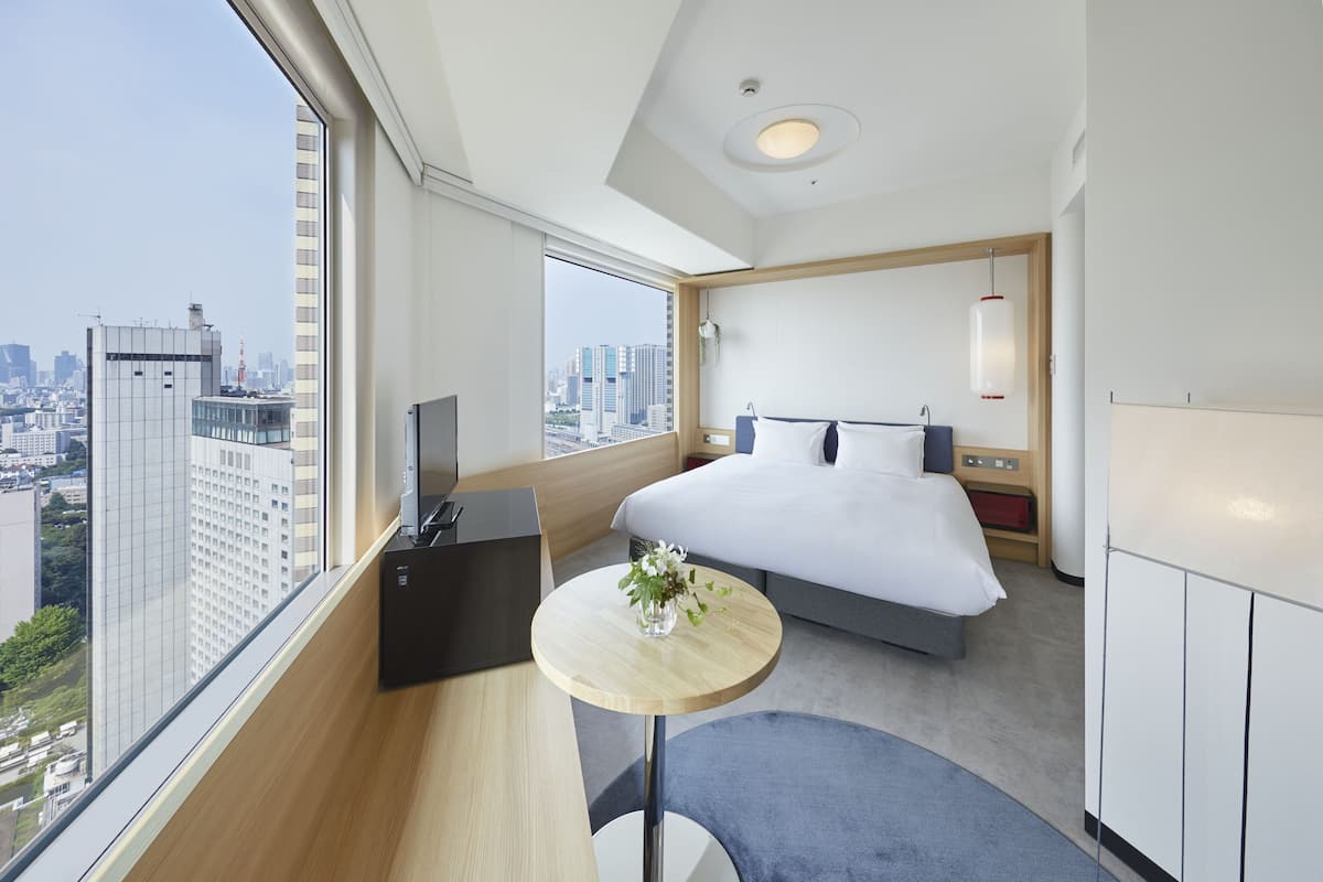 A modern hotel room with a large bed, a small round table with a vase of flowers, a TV, and a view of the cityscape.
