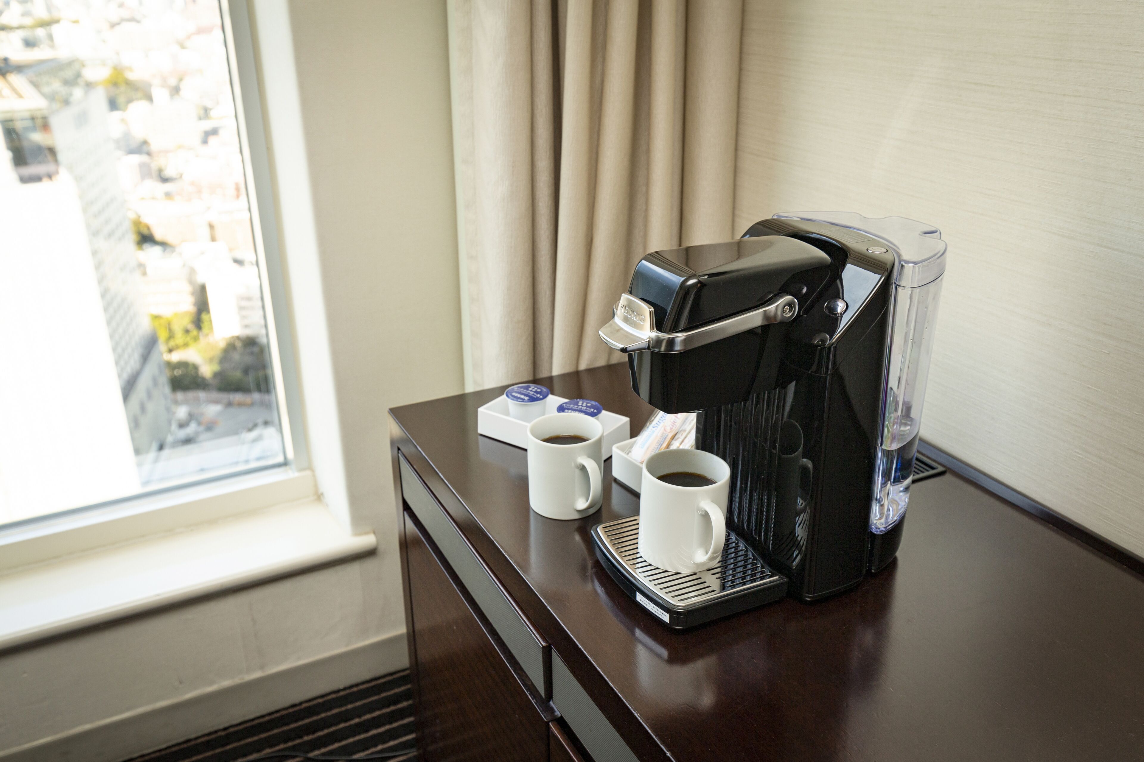 A coffee maker on a wooden table with two white cups and a box of coffee pods.