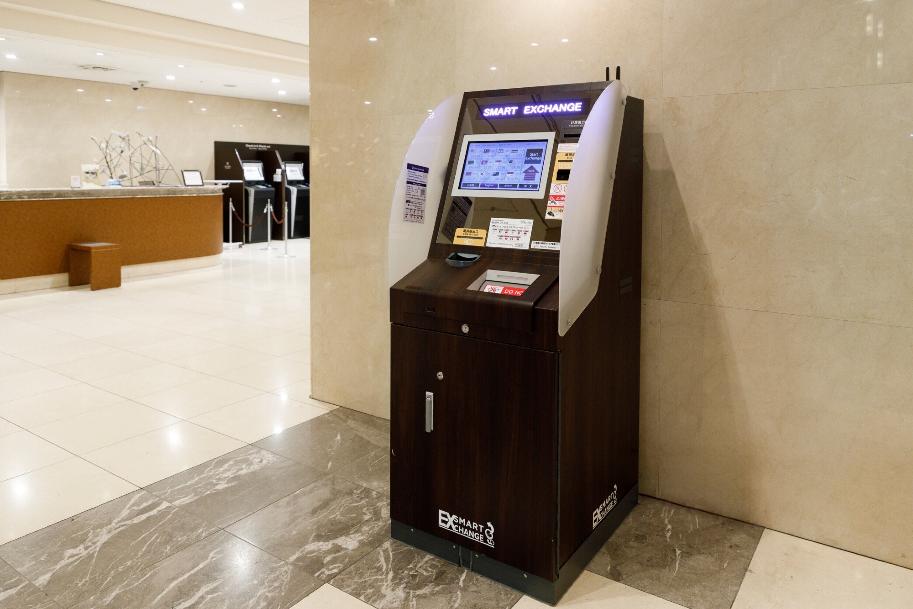 Un quiosco de intercambio automatizado en el vestíbulo de un hotel, con piso de mármol y un mostrador de recepción al fondo.