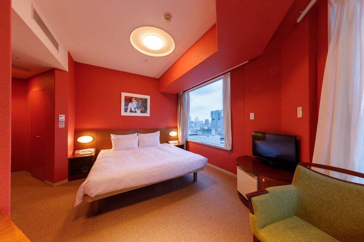 A hotel room with a bed, a television, a green armchair, a window with a city view, and red walls.